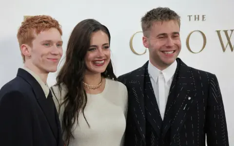 Skådespelare från "The Crown" ler glatt. En man med rött hår i mörk kavaj, en kvinna i ljus tröja och en man i mönstrad kavaj.