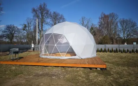 Nowoczesny glamping nad jeziorem. Biała kopuła na drewnianym tarasie, obok grill. Idealne miejsce na relaks.