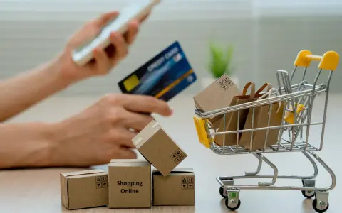 Manos sostienen un teléfono y tarjeta de crédito, junto a un carrito de compras con cajas y bolsas. El modelo 369 representa la facilidad de comprar en línea.