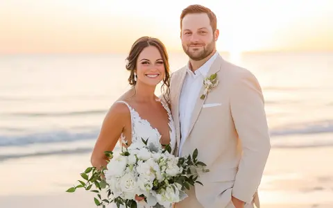 Groom in a tan suit, perfect for what to wear to beach wedding male, smiles with bride holding bouquet at sunset.
