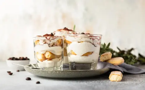 Pyszne desery włoskie: tiramisu w szklankach, udekorowane kakao i kawą. Obok kawałki biszkoptów i ziarna kawy.