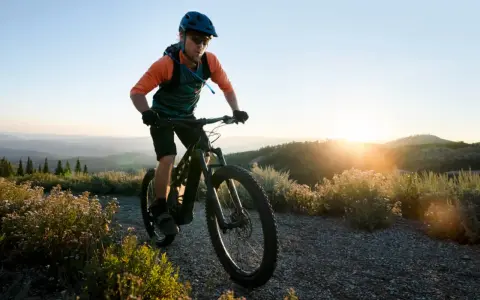Kolarz w kasku i okularach jedzie rowerem po górskiej ścieżce. Słońce zachodzi za wzgórzami, tworząc piękne światło. To idealny moment na maraton MTB.
