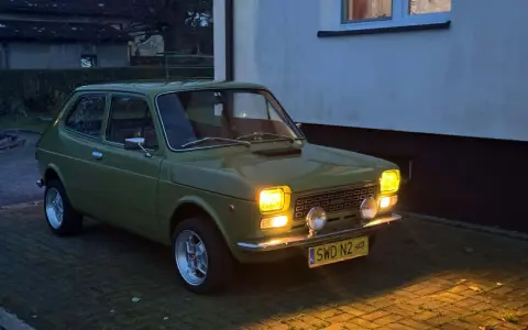 Zielony Fiat 127 z lat 70. stoi na podjeździe, jego światła rozświetlają zmierzch.