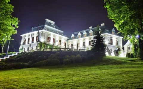 Elegancki hotel w lesie nocą, oświetlony blaskiem okien i zewnętrznych lamp. Otoczony zielenią, zaprasza do odpoczynku.
