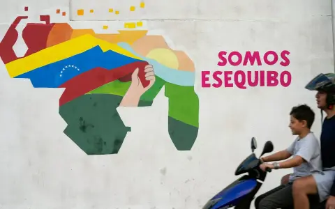 Mural przedstawiający mapę Wenezueli z flagą i hasłem "Somos Esequibo". Dwóch chłopców na skuterze przejeżdża obok.