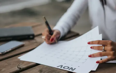 Une main avec des ongles rouges écrit sur un calendrier, planifiant peut-être la date de mariage. Un téléphone et un carnet sont à proximité.