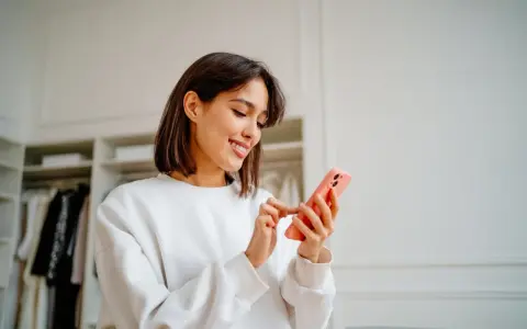 Uśmiechnięta kobieta przegląda telefon w swojej garderobie. Jej gotowy biogram czeka na publikację.