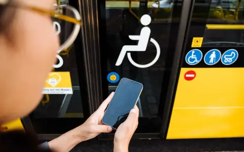 Osoba trzyma telefon przed drzwiami autobusu z symbolem wózka inwalidzkiego. To ułatwia podróżowanie, gdy masz legitymację osoby niepełnosprawnej.