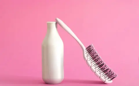 Eine weiße Flasche, die wie ein **collagen shampoo** aussieht, lehnt an einer Haarbürste auf rosa Hintergrund.