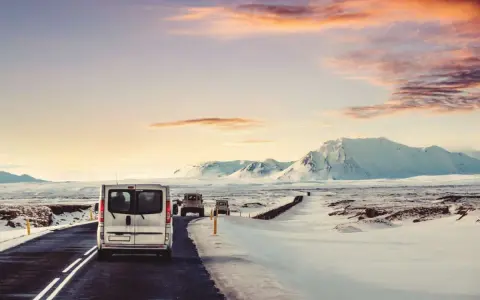 A silver van leads a convoy of vehicles driving in Iceland in December, past snow-covered mountains under a dramatic sunset sky.