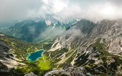 Blick vom Drachenkopf auf einen türkisfarbenen Bergsee, umgeben von grünen Hängen und felsigen Gipfeln, Wolken ziehen über die Landschaft.