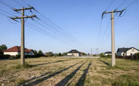 Linia średniego napięcia na działce, z domami w tle. Słupy energetyczne przecinają niebo, a ich cienie padają na suchą trawę.