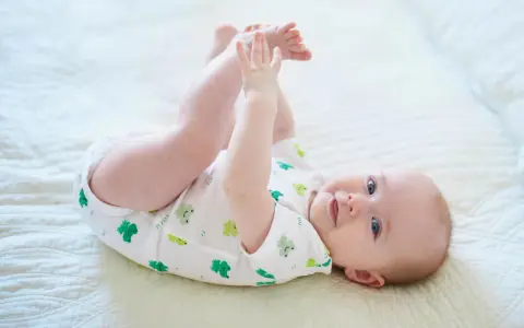 Ein Baby im 6. Lebensmonat liegt auf dem Rücken und spielt mit seinen Füßen. Es trägt einen weißen Body mit grünen Fröschen.