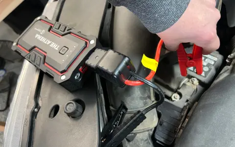 A hand connects red jumper cables from "The Athena" lithium jump starter to a car battery, demonstrating how lithium jump starters work by providing a temporary power boost.