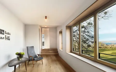 Interior moderno con suelo de madera, sillón gris y gran ventanal. El marco de madera de la ventana, que parece recién restaurado, enmarca un paisaje exterior.