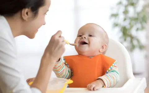 Mama karmi uśmiechniętego niemowlaka łyżeczką. To idealny przykład, co na śniadanie dla niemowlaka – zdrowo i radośnie!