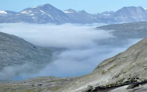 Moln sveper genom dalarna i Padjelanta nationalpark, med snöklädda bergstoppar som reser sig mot en klarblå himmel.