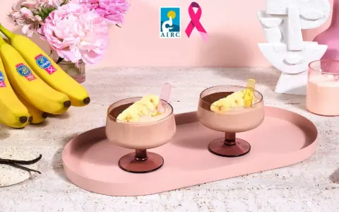 Due coppe di cremoso gelato alla banana, guarnite con fettine di banana e cacao, su un vassoio rosa. Banane fresche e fiori rosa sullo sfondo.