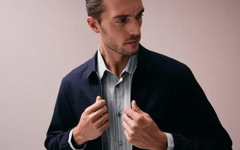 Ein Mann mit Bart und kurz geschnittenem Haar, der eine dunkelblaue Jacke über einem gestreiften Hemd trägt. Er fasst nach dem Reißverschluss, als würde er seine Kleidung für das Hemden waschen vorbereiten.