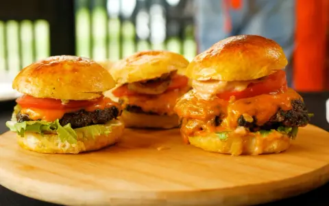Trzy soczyste burgery z roztopionym serem, pomidorem i sałatą na desce.