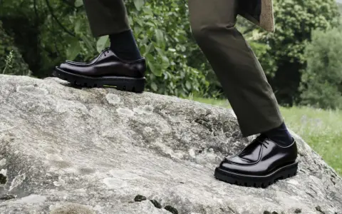 Scarpe oxford derby lucide su una roccia, con pantaloni verdi e sfondo verdeggiante.