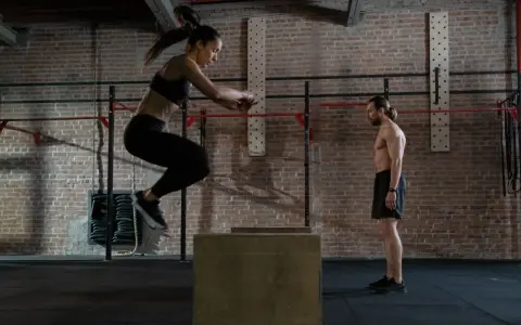 Frau springt auf eine Box, um ihre Kondition aufzubauen. Mann beobachtet sie im Fitnessstudio.