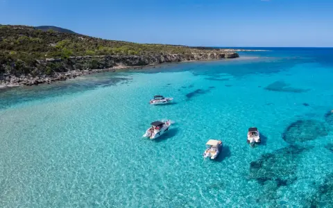 Traumhafter zypern strand mit kristallklarem Wasser und vier Booten, die vor der felsigen Küste ankern.
