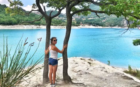 Donna sorridente abbraccia un albero con un lago turchese sullo sfondo. Una perfetta gita fuori porta nelle Marche.