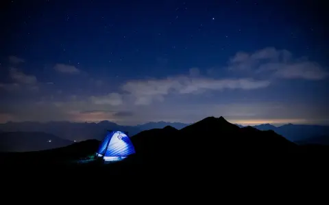 Une tente bleue s'illumine sous un ciel étoilé, témoin silencieux d'un trek inoubliable au cœur des montagnes.