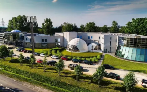 Nowoczesne polskie hotele niezależne z kopułami i przeszklonymi fasadami, otoczone zielenią i parkingiem pełnym samochodów.