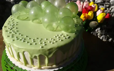 Zielony tort udekorowany koralikami i przezroczystymi, zielonymi balonami z żelatyny. Obok bukiet kolorowych tulipanów.