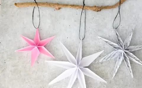 Drei Papiersterne, die Kinder falten könnten, hängen an einem Ast. Einer ist rosa, die anderen weiß und mit Noten bedruckt.