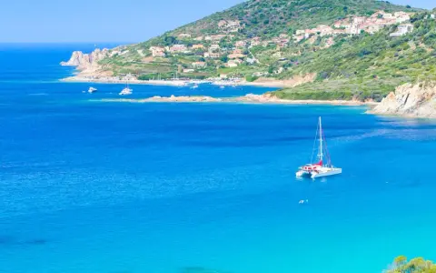Un catamaran blanc avec des accents rouges navigue sur des eaux bleues cristallines, près d'une côte verdoyante et d'un village pittoresque.