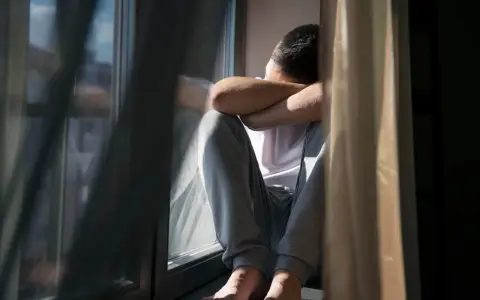 Mężczyzna siedzi przy oknie, obejmując się ramionami, pogrążony w myślach.