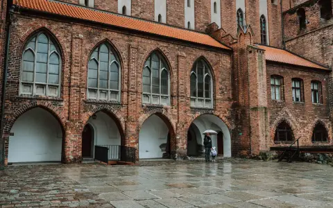 Zamek w Kwidzynie, z ceglanymi arkadami i gotyckimi oknami, skrywa tajemnice. Dwoje ludzi z parasolem spaceruje po mokrym dziedzińcu.