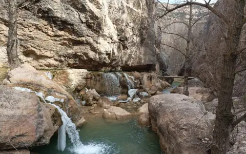 Cascada y río en un cañón rocoso, con hielo en las rocas. Un puente de madera cruza el río, evocando las pasarelas de Valloré.