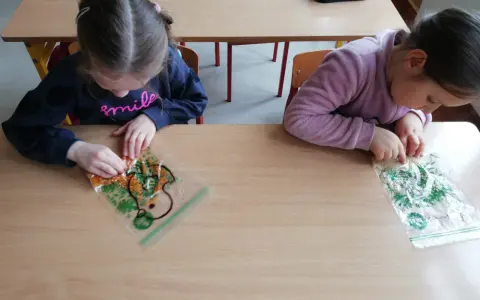 Dwie dziewczynki bawią się materiałami sensorycznymi w woreczkach na drewnianym stole.