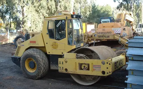 Żółty walec drogowy BOMAG BW 213D-3 gotowy do budowy drogi. W tle koparka Samsung Caterpillar.