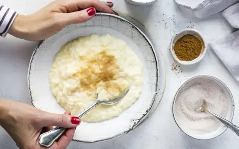 Cremiger Milchreis mit Vanillepudding, bestreut mit Zimt. Eine Hand mit roten Nägeln hält einen Löffel bereit.