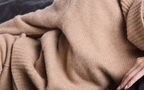 Eine Frau in einem beigen Kaschmirpullover liegt entspannt auf einem dunklen Sofa. Das Kaschmir waschen ist wichtig für die Pflege.