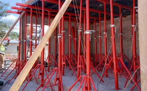 Construcción con varios tipos de puntales rojos sosteniendo una estructura. Un trabajador supervisa.