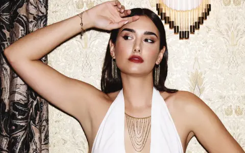 Mujer con pendientes media luna dorados, collar en cascada y pulsera de corazones, posando con un vestido blanco halter.