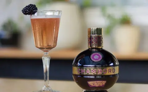 Elegancki kieliszek z różowym drinkiem i jeżyną obok butelki likieru Chambord.