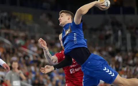 Magnus Landin Jacobsen w skoku do rzutu piłką ręczną, w niebieskim stroju, z widownią w tle.