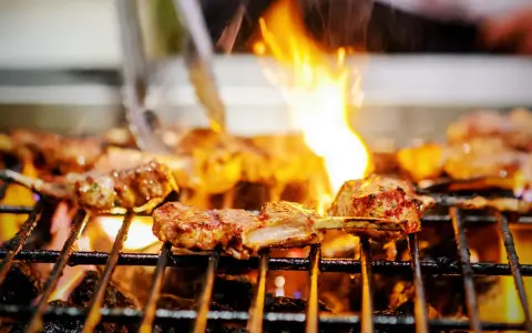 Marinierte Lammkoteletts brutzeln auf dem Grill. Flammen züngeln über dem Fleisch, das perfekt gegrillt wird.