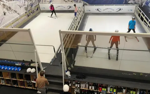 Ludzie ćwiczą jazdę na nartach na sztucznym stoku narciarskim w Ski Arenie Wrocław. Widać sprzęt narciarski i buty narciarskie w schowkach.