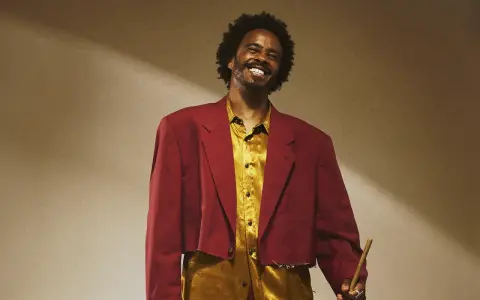 Un percussionniste célèbre, souriant, porte une veste courte bordeaux et une chemise dorée, tenant une baguette.