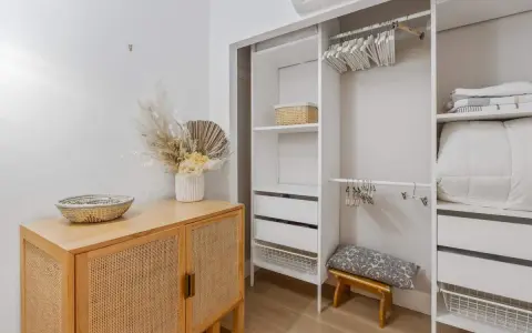 Un armario blanco organizado, listo para limpiar armarios por dentro. Cestas, ropa de cama y un jarrón con flores secas decoran el espacio.