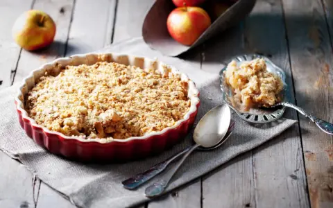 Un délicieux crumble pomme individuel, doré et croustillant, prêt à être dégusté. Des pommes fraîches sont visibles en arrière-plan.
