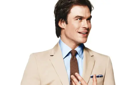 A man in a tan suit and blue shirt smiles, adjusting his tie.